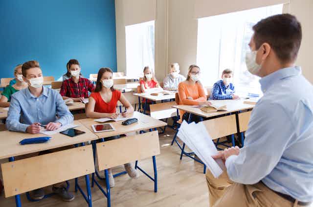 Masked students in classroom listening to masked teacher