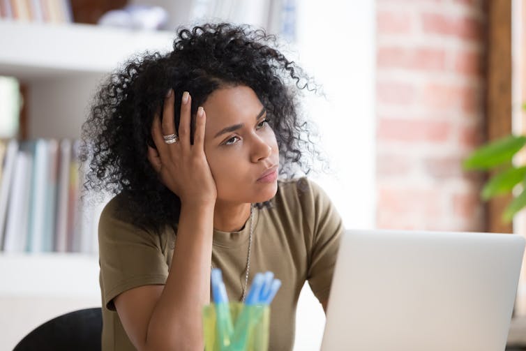 Black woman sits at a desk with her laptop, head in her hands, looking exasperated.