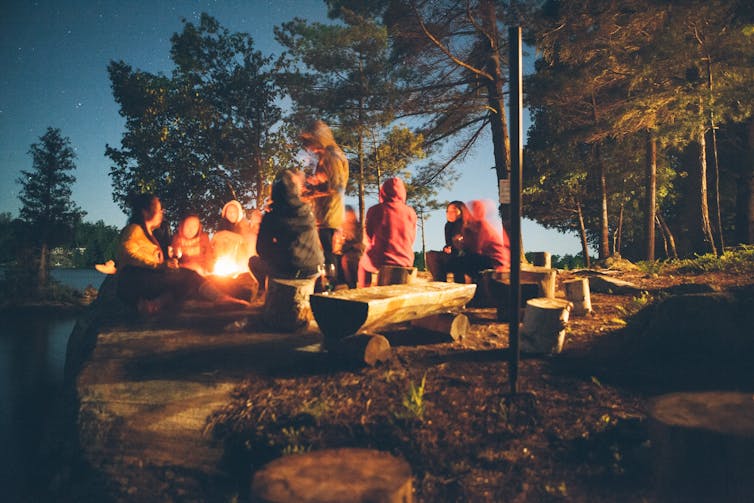 Group of people sit in forest near bonfire.