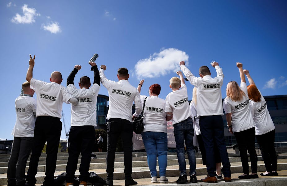 A line of 9 people, seen from the back, most with fists or peace fingers in the air. They are all wearing white shirts with text reading "the truth is free"