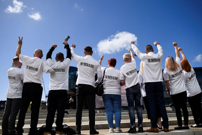 A line of 9 people, seen from the back, most with fists or peace fingers in the air. They are all wearing white shirts with text reading "the truth is free"