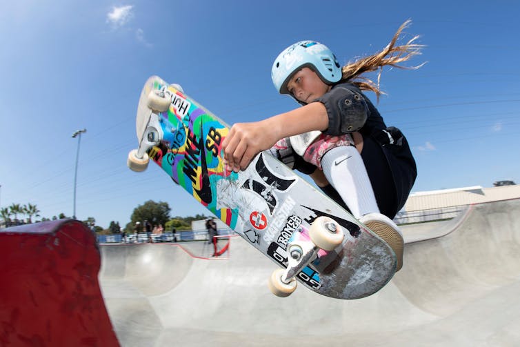 A young girl in a helmet, black sports clothes and white socks and trainers does a jump on her skateboard
