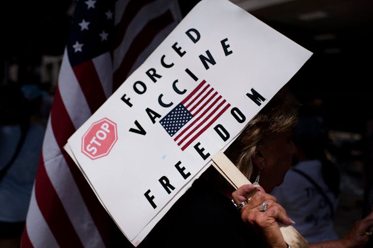 Anti-vaccine rally protester holds a sign saying 'Stop Forced Vaccine' above an American flag.