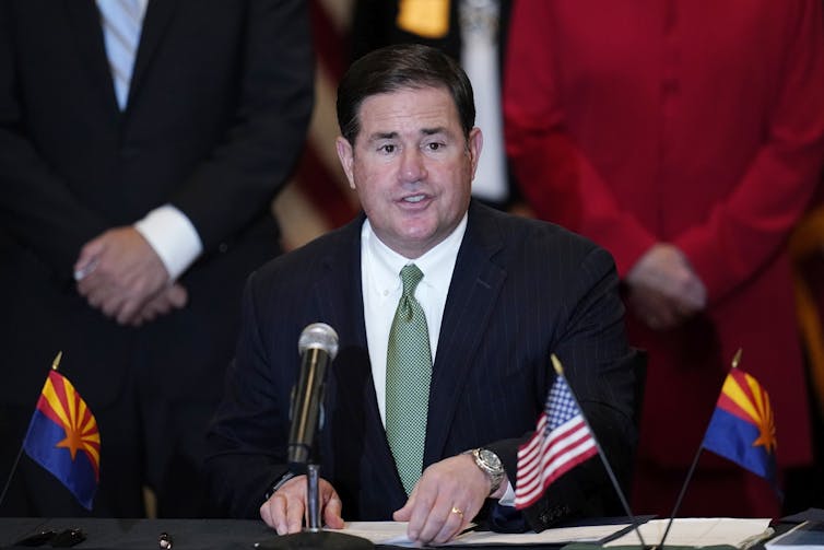 Arizona Gov. Doug Ducey in a black blazer and green tie in front of a microphone.