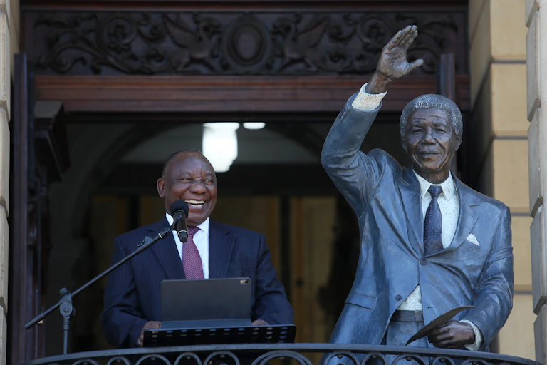 A man wearing a tie and suit smiles while standing at a podium with a mircophone next to a statue of a man
