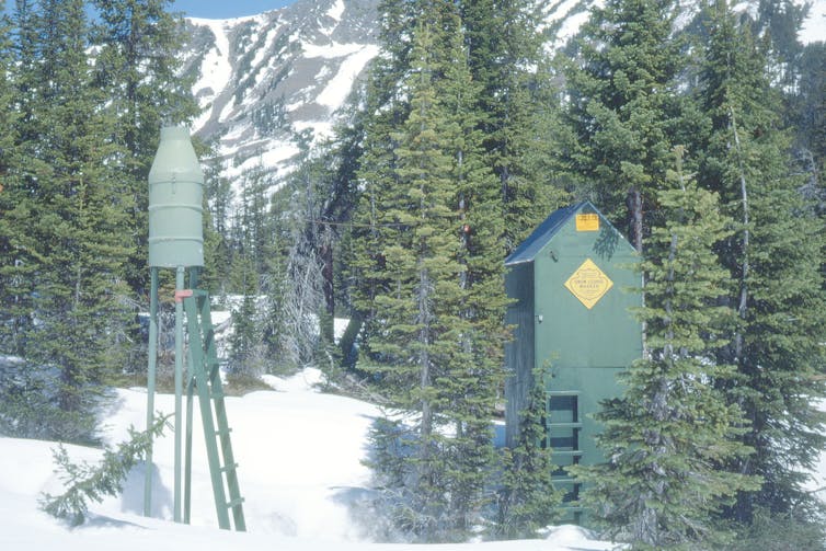 A small green metal tower and green wooden box in a snowy mountain forest.