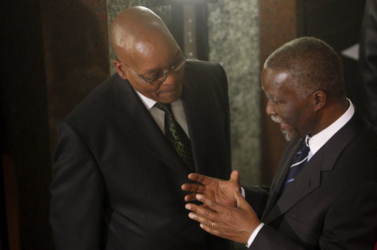 A man wearing spectacles, a suit and tei listens attentively to another.