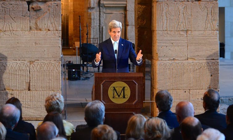 John Kerry giving speech at lectern in front of Syrian artifacts.