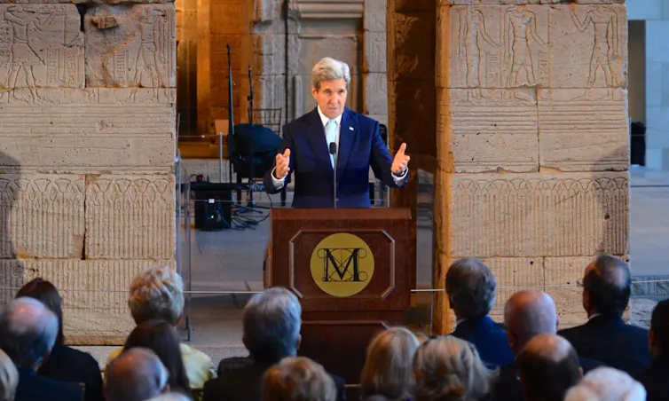 John Kerry giving speech at lectern in front of Syrian artifacts.