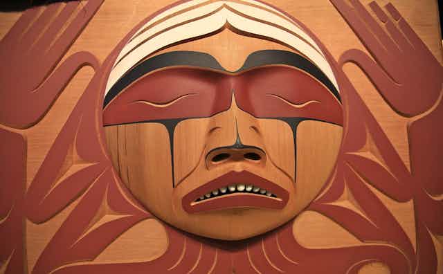 Carving of a round crying face surrounded by palms extended upwards.