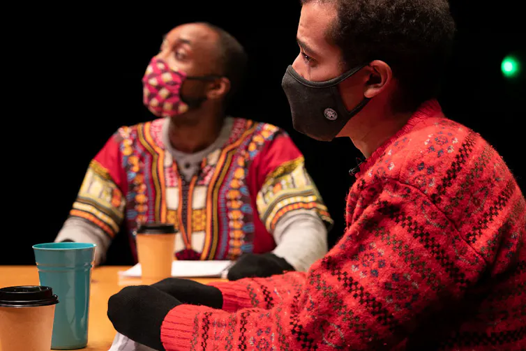 Two actors sitting at a table with coffee cups on a stage.
