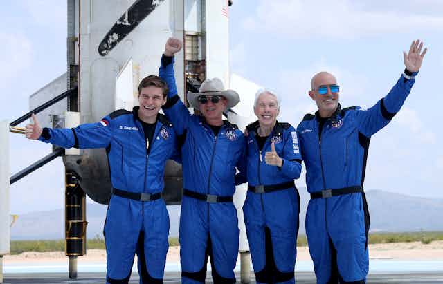 Four white people wearing space suits take a photo in front of a spaceship.