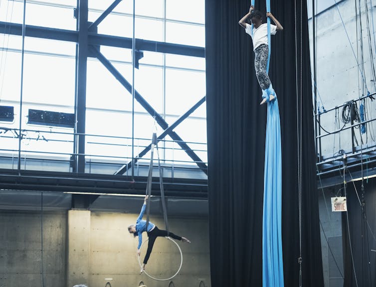 Two children performing aerial circus arts: on climbing a blue rope and one on a suspended hoop