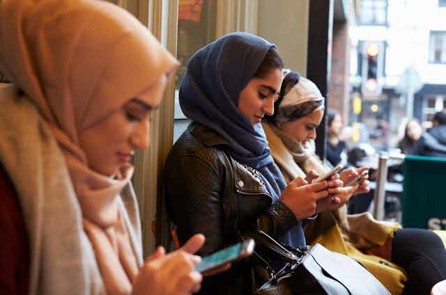 Three women wearing hijabs sitting side by side outside a shop and texting