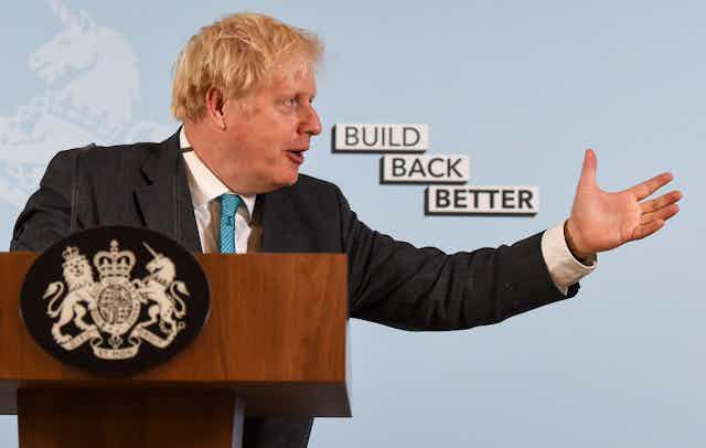 Boris Johnson gestures while speaking at a podium, with a background reading "Build Back Better." His arm is extended towards the text.