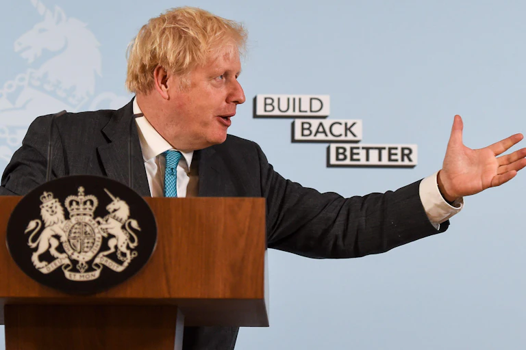 Boris Johnson gestures while speaking at a podium, with a background reading "Build Back Better." His arm is extended towards the text.