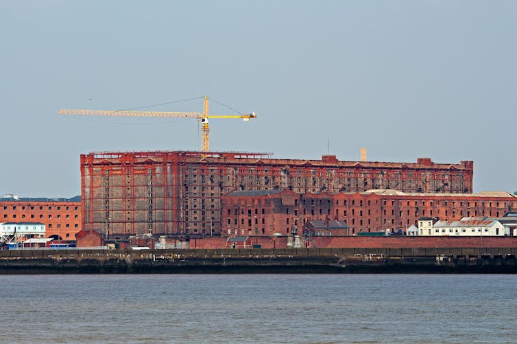 Cranes and scaffolding surround a large red-brick warehouse seen from across the water