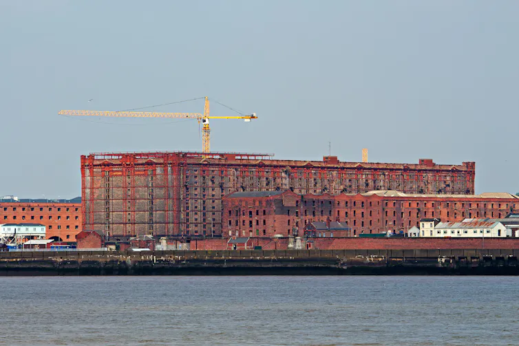 Cranes and scaffolding surround a large red-brick warehouse seen from across the water
