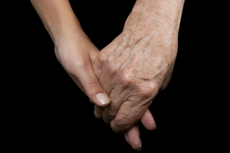 A young person's hand and an old person's hand clasped against a black background.