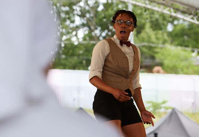 An actor on a stage in glassses, a bow tie, and shorts shows a surprised face with mouth agape.