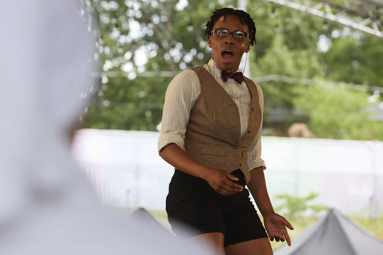 An actor on a stage in glassses, a bow tie, and shorts shows a surprised face with mouth agape.