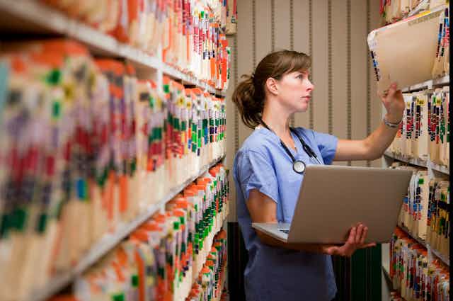 Health care provider holding laptop pulls out a medical record from a shelf.