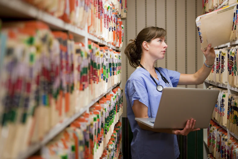 Health care provider holding laptop pulls out a medical record from a shelf.