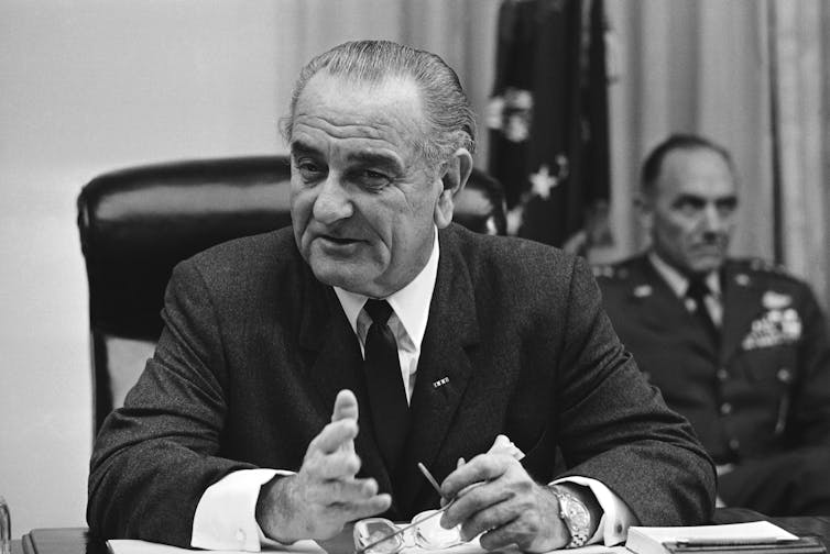 President Lyndon Johnson, sitting at his desk in a suit, tie and white shirt.