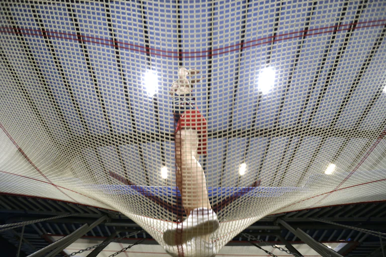 A male gymnast is seen through the holes of a trampoline as he bounces on it.