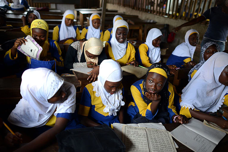A classroom with young female learners, books open in front of them, wearing hijabs.