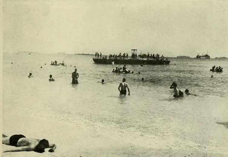 People swimming in a lagoon, with some sunbathing on a beach, ships in the background.