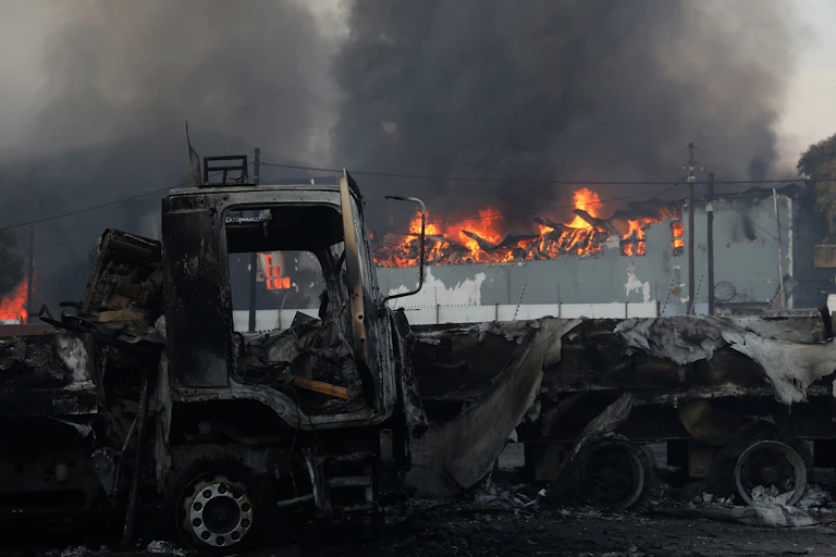 A business building is engulfed in flames next to burnt trucks.