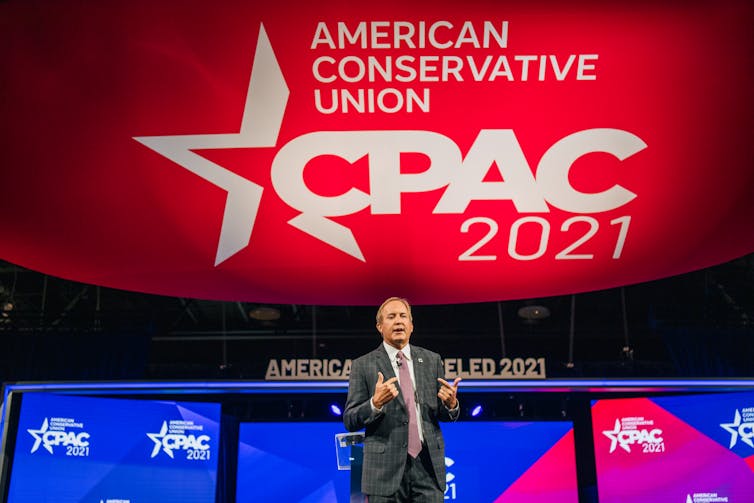 Paxton speaks, standing on a stage in front of a big CPAC 2021 backdrop