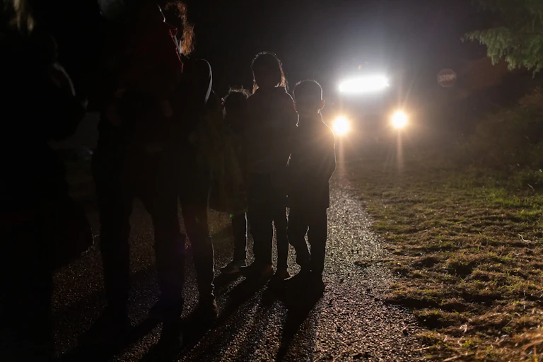 Three children stand on a street night, backlit by car headlights