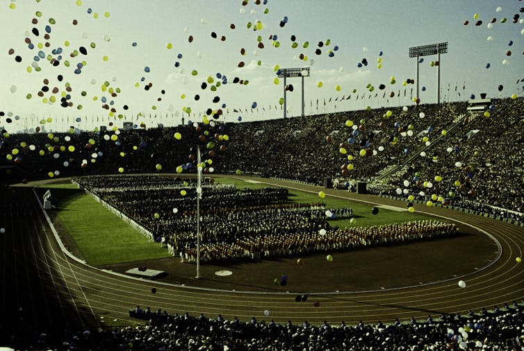 balões voam sobre atletas olímpicos e espectadores durante a cerimônia de abertura dos Jogos Olímpicos de Verão de 1964 no Estádio Nacional de Tóquio.