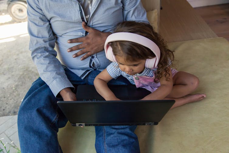 A young Aboriginal child is playing on a laptop with headphones on, next to an Aboriginal adult.