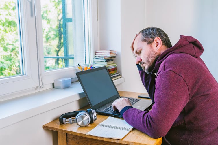 Frustrated looking man sits at laptop next to window