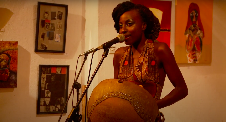 A glamorous woman in African-print attire holds a traditional musical instrument and sings into a microphone, art on the walls behind her.