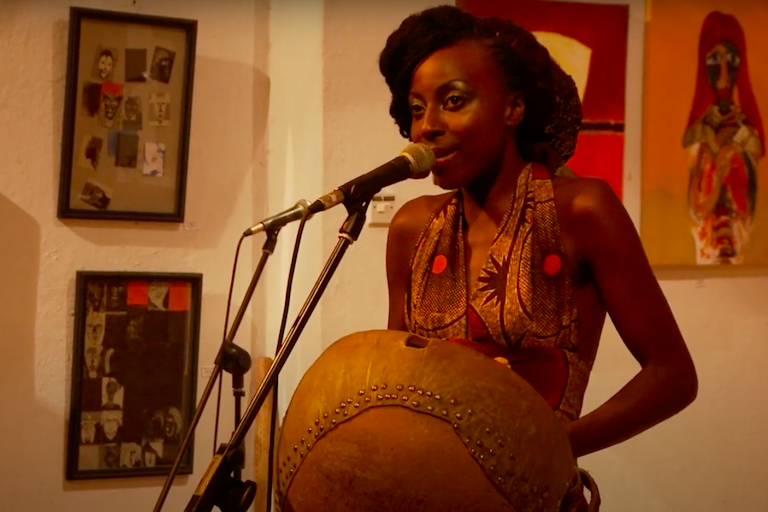 A glamorous woman in African-print attire holds a traditional musical instrument and sings into a microphone, art on the walls behind her.