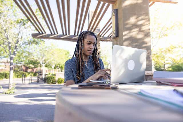 A queer student uses their laptop outside on a desk.