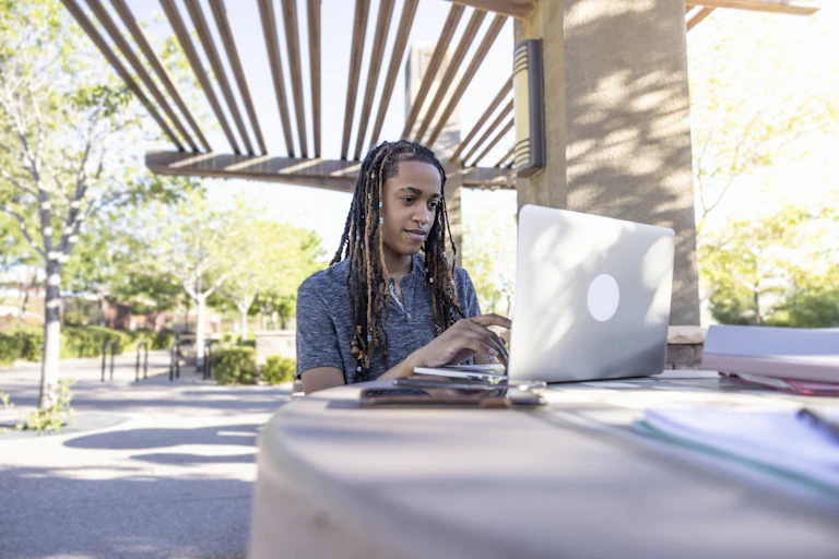 A queer student uses their laptop outside on a desk.