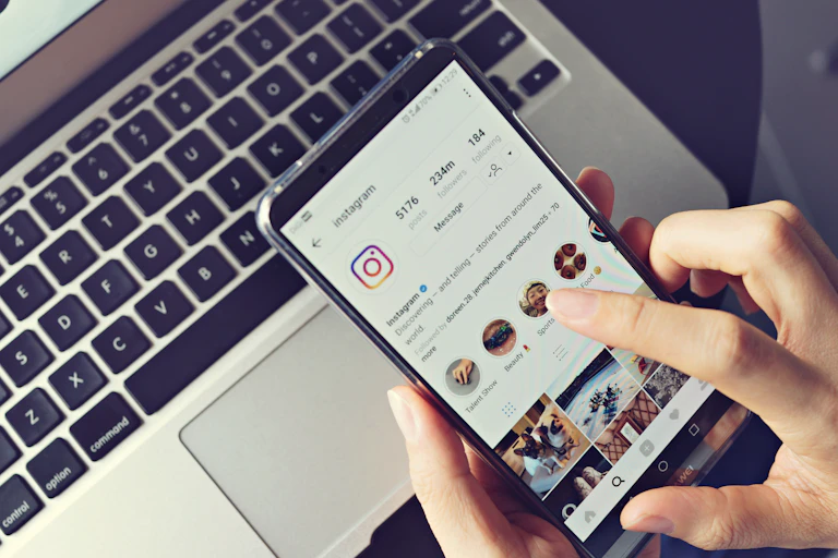 A manicured hand holds a phone showing Instagram, an Apple laptop keyboard is visible in the background
