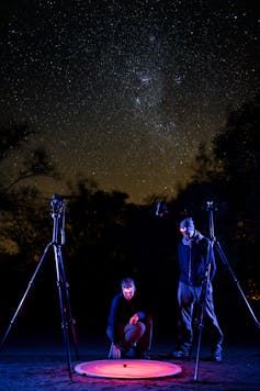 Two people, both in dark clothing, are standing alongside two cameras. One is squatting, one is standing. The sky is full of stars, and the people are gazing at a small object on a lit-up pink circle on the ground