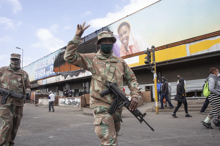 A man wearing camouflage uniform and carrying a gun raises two fingers in the peace sign while walking along a street, while another man in the same uniform follows.