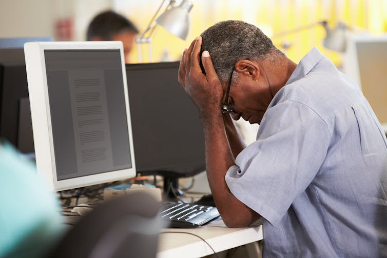 Older man holds head in front of a computer.