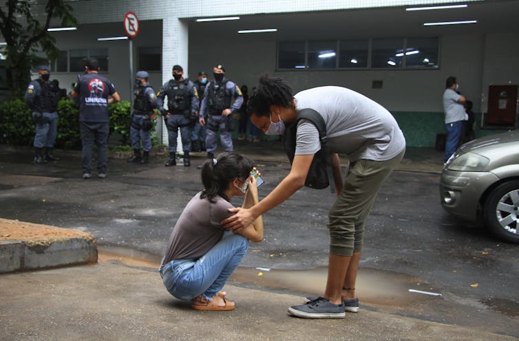 Woman is on knees crying outside of a building, a masked woman comforts her