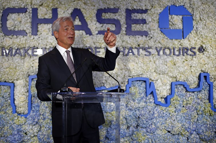 Jamie Dimon raises his left hand and points his index finger in a gesture as he stands in front of a glass lectern with the Chase logo behind him and an outline of a city in blue.