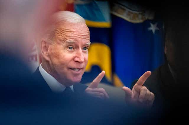 President Biden gestures with both hands and index fingers pointing as his face is framed through indistinct shapes of people in the foreground
