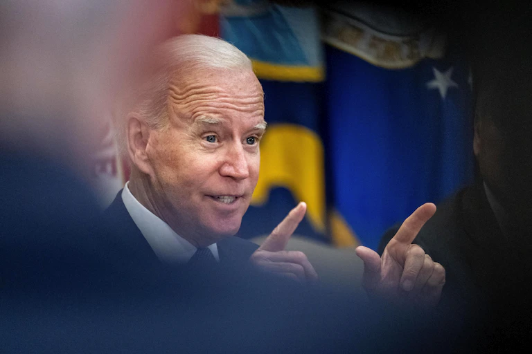 President Biden gestures with both hands and index fingers pointing as his face is framed through indistinct shapes of people in the foreground