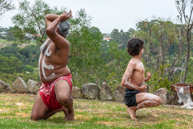 Aboriginal half-naked men with white stripe on his body and groin cloth strike defiant poses while kneeling on the ground.